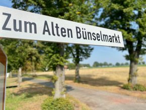 Ein Straßenschild mit dem Straßennamen "Zum Alten Bünselmark". Im Hintergrund ist leicht verschwommen das Baugebiet zu erkennen. 