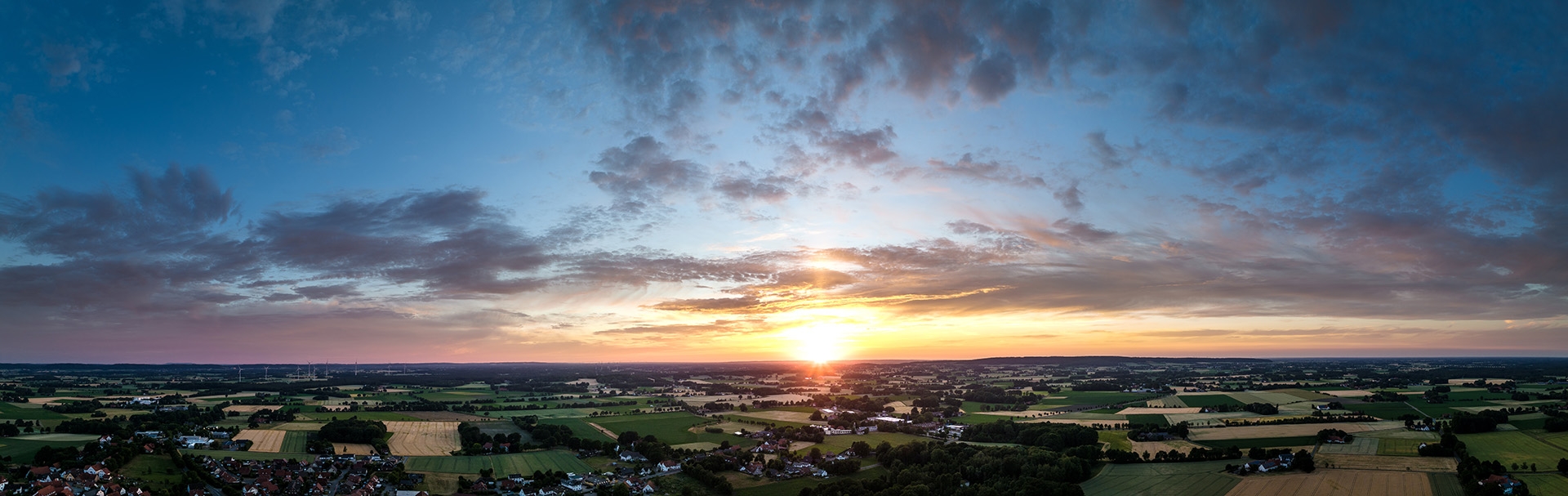 Weite Landschaft mit Feldern, Wiesen und kleinen Ortschaften aus der Vogelperspektive. Am Horizont geht die Sonne unter und färbt den Himmel in Gelb-, Orange- und Blautöne. Vereinzelte Wolken ziehen über die Landschaft, im Vordergrund sind Dächer, Bäume und landwirtschaftliche Flächen zu erkennen.