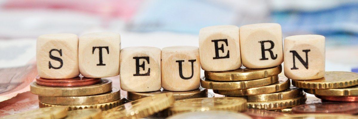 Holzwürfel, auf denen das Wort Steuern zu lesen ist, liegen auf einem Haufen Geldscheine und Münzen.