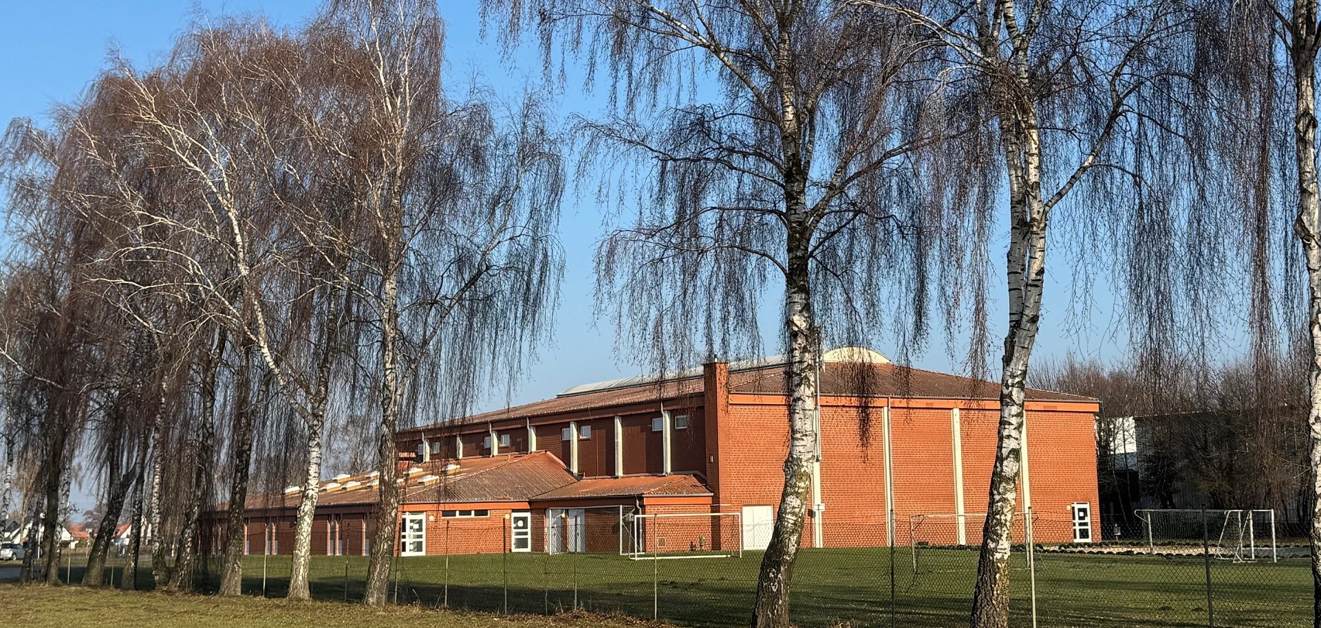 Rote Sporthalle aus Backstein mit flachem, leicht geneigtem Dach, aufgenommen an einem sonnigen Tag. Im Vordergrund stehen mehrere hohe, kahle Birken auf einer Wiese. Die Halle liegt hinter einem niedrigen Zaun, seitlich sind Sportgeräte und Tore zu erkennen. Blauer Himmel ohne Wolken.
