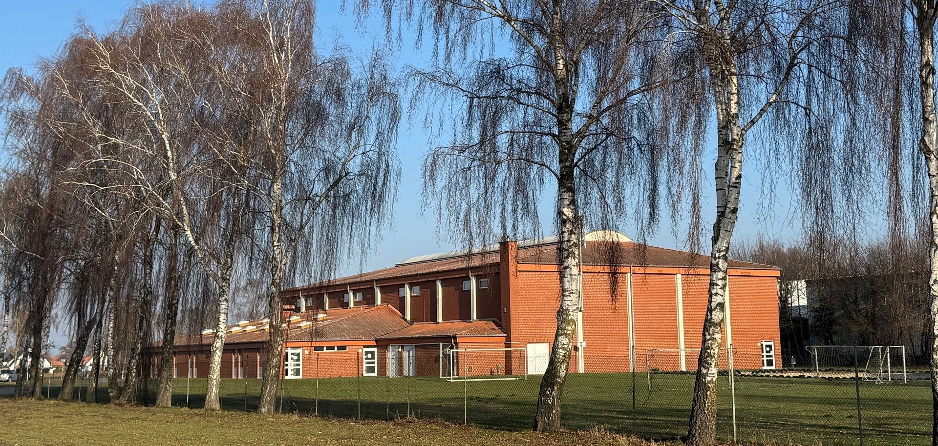 Rote Sporthalle aus Backstein mit flachem, leicht geneigtem Dach, aufgenommen an einem sonnigen Tag. Im Vordergrund stehen mehrere hohe, kahle Birken auf einer Wiese. Die Halle liegt hinter einem niedrigen Zaun, seitlich sind Sportgeräte und Tore zu erkennen. Blauer Himmel ohne Wolken.