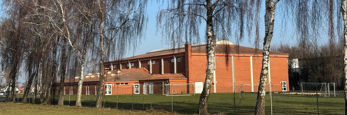 Sporthalle Dielingen 2026 Rote Sporthalle aus Backstein mit flachem, leicht geneigtem Dach, aufgenommen an einem sonnigen Tag. Im Vordergrund stehen mehrere hohe, kahle Birken auf einer Wiese. Die Halle liegt hinter einem niedrigen Zaun, seitlich sind Sportgeräte und Tore zu erkennen. Blauer Himmel ohne Wolken.
