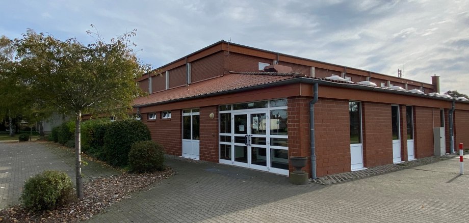 Außenansicht einer eingeschossigen Sporthalle aus rotem Klinker. Im Vordergrund befindet sich ein gepflasterter, ebener Zugangsbereich. Zentral sind breite, verglaste Doppeltüren mit weißen Rahmen zu sehen, daneben weitere Türen und Fenster. Das Gebäude hat ein flach geneigtes Dach mit Dachrinnen. Links vom Eingang stehen Bäume und Sträucher, rechts ist ein freier Platz. Der Himmel ist leicht bewölkt.