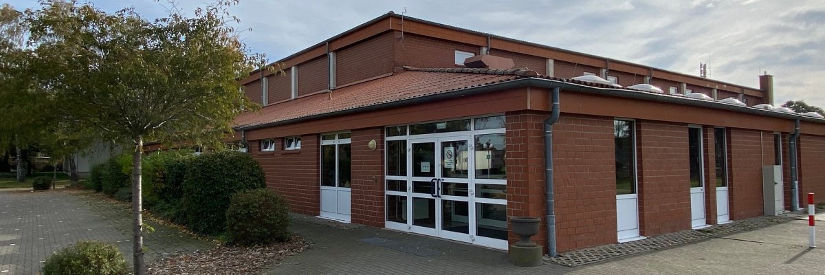 Außenansicht einer eingeschossigen Sporthalle aus rotem Klinker. Im Vordergrund befindet sich ein gepflasterter, ebener Zugangsbereich. Zentral sind breite, verglaste Doppeltüren mit weißen Rahmen zu sehen, daneben weitere Türen und Fenster. Das Gebäude hat ein flach geneigtes Dach mit Dachrinnen. Links vom Eingang stehen Bäume und Sträucher, rechts ist ein freier Platz. Der Himmel ist leicht bewölkt.