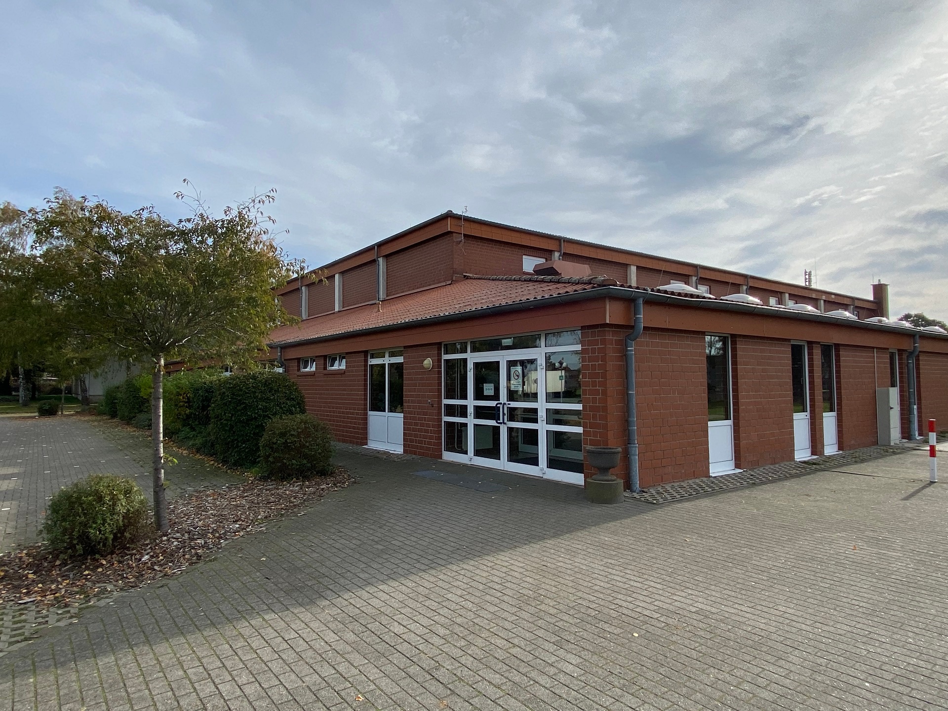 Außenansicht einer eingeschossigen Sporthalle aus rotem Klinker. Im Vordergrund befindet sich ein gepflasterter, ebener Zugangsbereich. Zentral sind breite, verglaste Doppeltüren mit weißen Rahmen zu sehen, daneben weitere Türen und Fenster. Das Gebäude hat ein flach geneigtes Dach mit Dachrinnen. Links vom Eingang stehen Bäume und Sträucher, rechts ist ein freier Platz. Der Himmel ist leicht bewölkt.