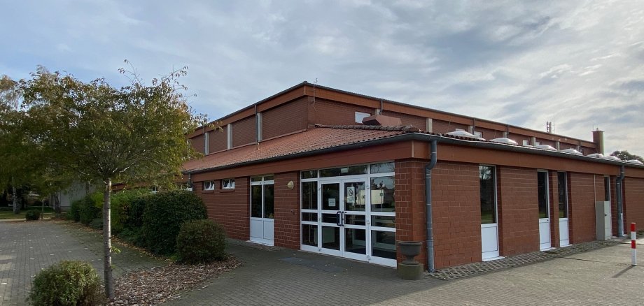 Außenansicht einer eingeschossigen Sporthalle aus rotem Klinker. Im Vordergrund befindet sich ein gepflasterter, ebener Zugangsbereich. Zentral sind breite, verglaste Doppeltüren mit weißen Rahmen zu sehen, daneben weitere Türen und Fenster. Das Gebäude hat ein flach geneigtes Dach mit Dachrinnen. Links vom Eingang stehen Bäume und Sträucher, rechts ist ein freier Platz. Der Himmel ist leicht bewölkt.