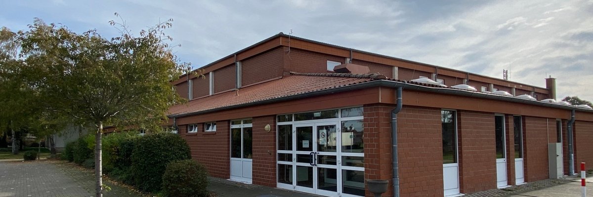 Sporthalle Dielingen Außenansicht einer eingeschossigen Sporthalle aus rotem Klinker. Im Vordergrund befindet sich ein gepflasterter, ebener Zugangsbereich. Zentral sind breite, verglaste Doppeltüren mit weißen Rahmen zu sehen, daneben weitere Türen und Fenster. Das Gebäude hat ein flach geneigtes Dach mit Dachrinnen. Links vom Eingang stehen Bäume und Sträucher, rechts ist ein freier Platz. Der Himmel ist leicht bewölkt.