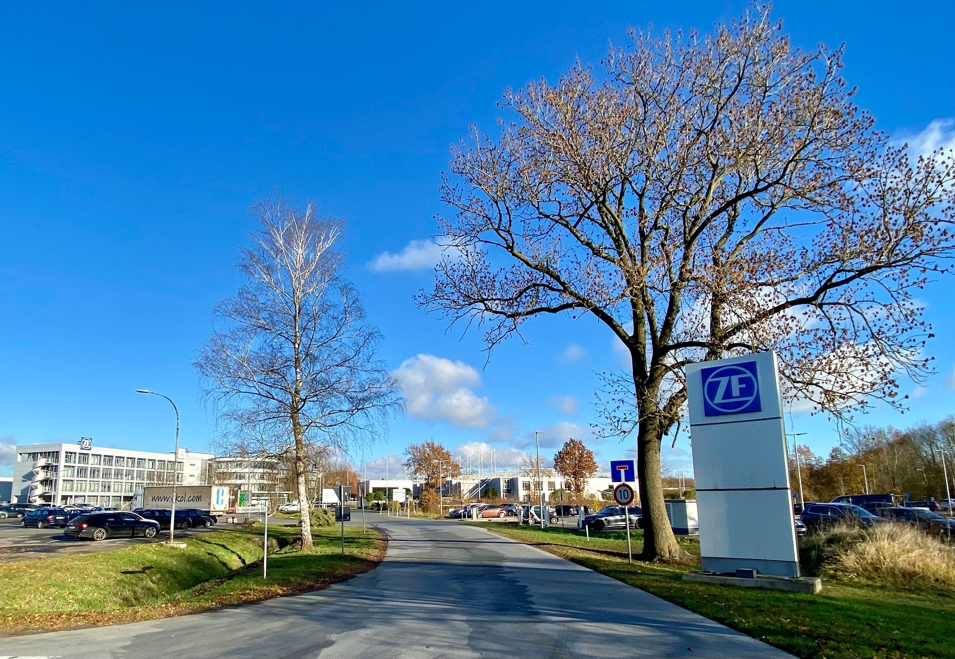 Zufahrtsstraße zu einem modernen Unternehmensgelände mit mehreren Gebäuden und Parkplätzen. Rechts im Vordergrund steht ein großes weißes Firmenschild mit blauem ZF-Logo neben einem hohen Laubbaum. Links säumen ein kahler Baum, geparkte Autos und ein Bürogebäude die Straße. Im Hintergrund sind weitere Betriebsgebäude und Parkflächen zu sehen. Der Himmel ist leuchtend blau mit wenigen weißen Wolken.
