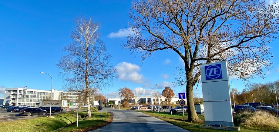 ZF Dielingen Zufahrtsstraße zu einem modernen Unternehmensgelände mit mehreren Gebäuden und Parkplätzen. Rechts im Vordergrund steht ein großes weißes Firmenschild mit blauem ZF-Logo neben einem hohen Laubbaum. Links säumen ein kahler Baum, geparkte Autos und ein Bürogebäude die Straße. Im Hintergrund sind weitere Betriebsgebäude und Parkflächen zu sehen. Der Himmel ist leuchtend blau mit wenigen weißen Wolken.