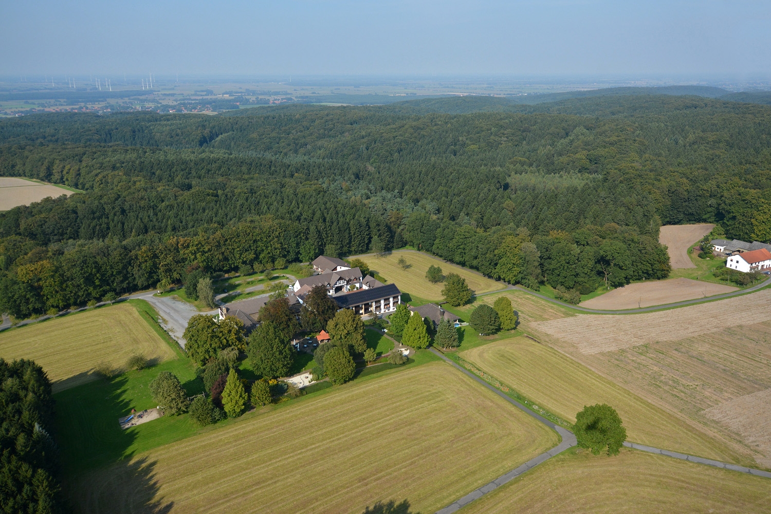 Luftaufnahme eines Landgasthofs mit mehreren Gebäuden, umgeben von Feldern, Wiesen und Waldgebieten; im Hintergrund sind Windräder und ein weiter Blick über die Landschaft zu sehen.