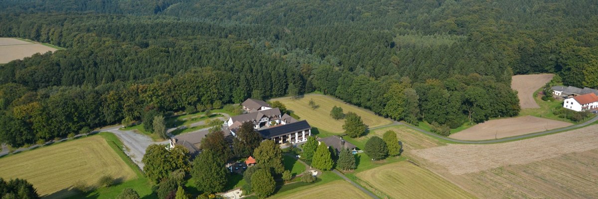 Luftaufnahme eines Landgasthofs mit mehreren Gebäuden, umgeben von Feldern, Wiesen und Waldgebieten; im Hintergrund sind Windräder und ein weiter Blick über die Landschaft zu sehen.