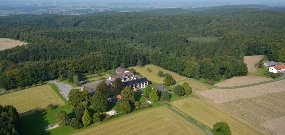 Luftaufnahme eines Landgasthofs mit mehreren Gebäuden, umgeben von Feldern, Wiesen und Waldgebieten; im Hintergrund sind Windräder und ein weiter Blick über die Landschaft zu sehen.