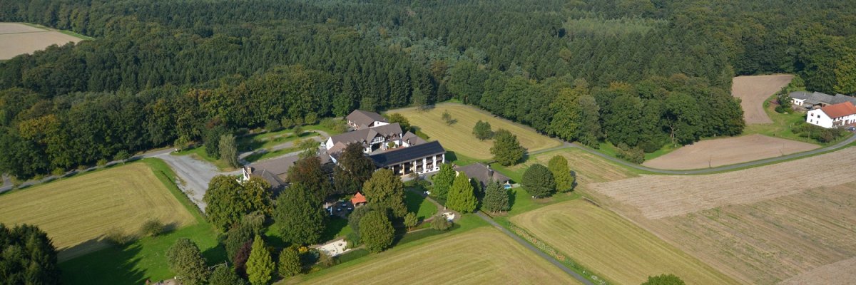 Wilhelmshöhe Luftaufnahme eines Landgasthofs mit mehreren Gebäuden, umgeben von Feldern, Wiesen und Waldgebieten; im Hintergrund sind Windräder und ein weiter Blick über die Landschaft zu sehen.