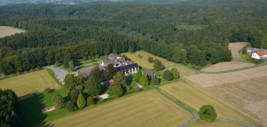 Wilhelmshöhe Luftaufnahme eines Landgasthofs mit mehreren Gebäuden, umgeben von Feldern, Wiesen und Waldgebieten; im Hintergrund sind Windräder und ein weiter Blick über die Landschaft zu sehen.