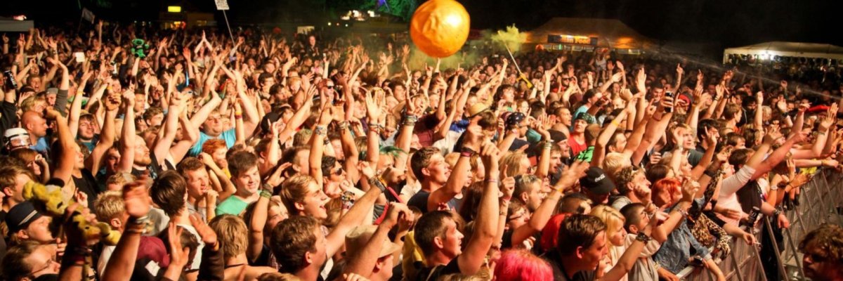 Viele Zuschauer, die im Bühnenlicht dichtgedrängt und mit nach oben gerissenen Armen vor einer Bühne stehen und mitsingen. 