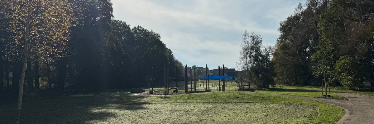 Weitläufige Parklandschaft an einem sonnigen Tag. Im Vordergrund liegt eine leicht feuchte, grünliche Rasenfläche. Rechts verläuft ein geschwungener, befestigter Weg. In der Mitte stehen mehrere Holzpfosten und Spiel- oder Bewegungsgeräte mit einem blauen Sonnensegel. Umgeben ist die Fläche von Bäumen mit teils herbstlichem Laub; im Hintergrund sind Häuser zu sehen. Der Himmel ist hellblau mit dünnen Wolken, die Sonne scheint von links oben.