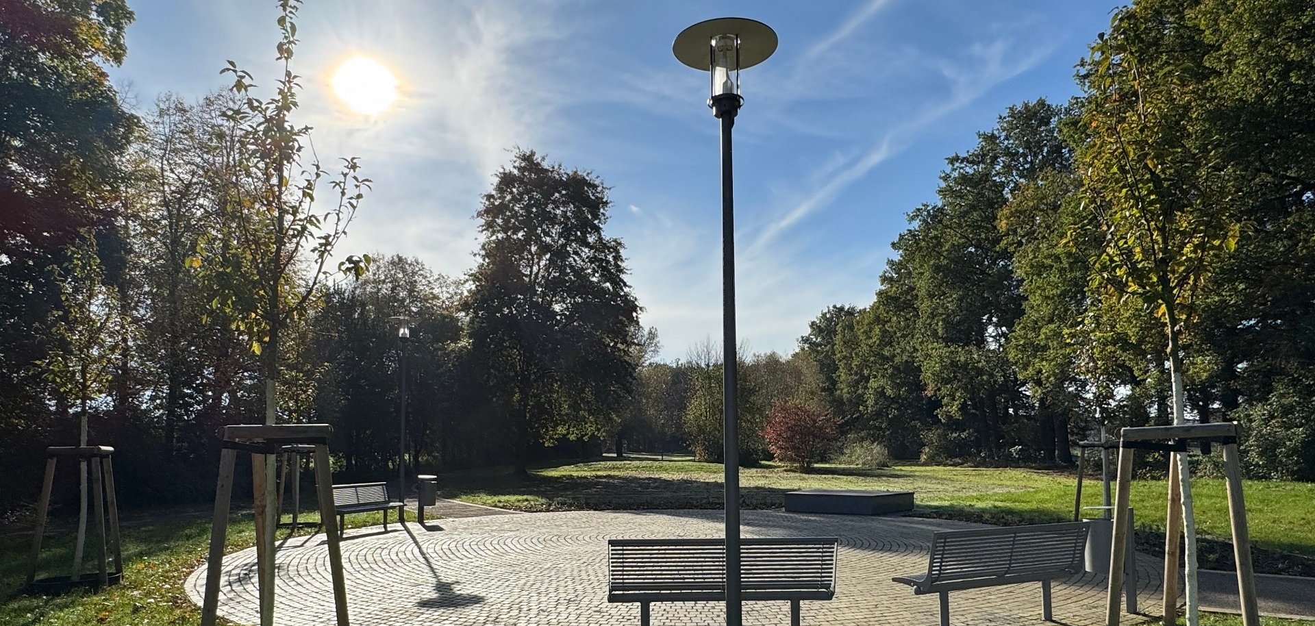 Runder, gepflasterter Platz in einem Park an einem sonnigen Tag. In der Mitte steht eine hohe schwarze Laterne. Mehrere Metallbänke sind kreisförmig um den Platz angeordnet. Im Vordergrund wächst grünes Gras; links und rechts stehen junge Bäume mit Holzstützen. Der Platz ist von Rasenflächen und dichtem Baumbestand umgeben. Die Sonne steht links am Himmel, der blau ist und von feinen Wolken durchzogen wird.