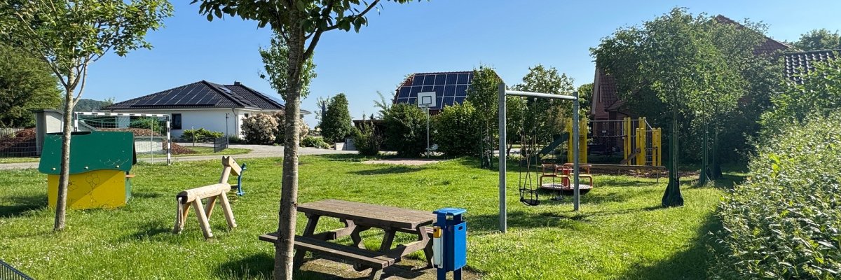 Spielplatz Heitkampsort Wehdem Sonniger Spielplatz mit viel Grünfläche, mehreren Bäumen und verschiedenen Spielgeräten. In der Mitte stehen ein Picknicktisch und ein Mülleimer, daneben eine Schaukel. Im Hintergrund befinden sich weitere Spielgeräte sowie Wohnhäuser mit Solaranlagen unter blauem Himmel.