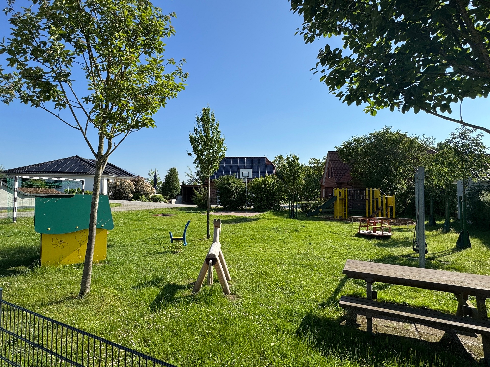 Sonniger Spielplatz mit viel Grünfläche, mehreren Bäumen und verschiedenen Spielgeräten. In der Mitte stehen ein Picknicktisch und ein Mülleimer, daneben eine Schaukel. Im Hintergrund befinden sich weitere Spielgeräte sowie Wohnhäuser mit Solaranlagen unter blauem Himmel.