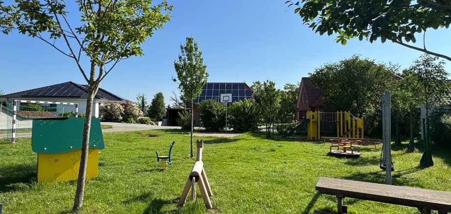 Spielplatz Heitkampsort Wehdem Sonniger Spielplatz mit viel Grünfläche, mehreren Bäumen und verschiedenen Spielgeräten. In der Mitte stehen ein Picknicktisch und ein Mülleimer, daneben eine Schaukel. Im Hintergrund befinden sich weitere Spielgeräte sowie Wohnhäuser mit Solaranlagen unter blauem Himmel.