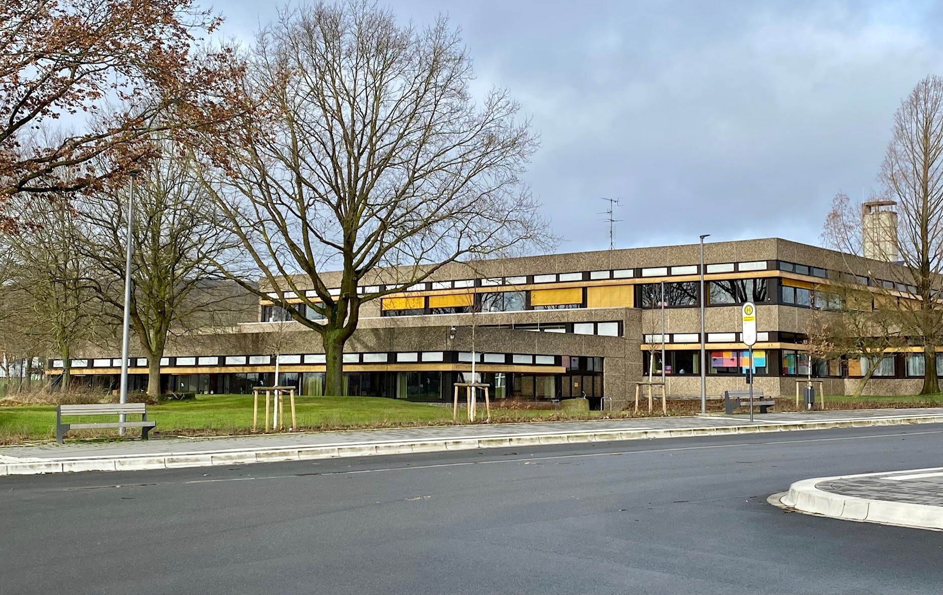 Außenansicht eines zweigeschossigen, langgestreckten Gebäudes mit dunkler Fassade und Fensterbändern. Im Vordergrund verlaufen eine Straße, ein Gehweg mit Sitzbank und eine Bushaltestelle. Mehrere kahle Bäume stehen vor dem Gebäude.