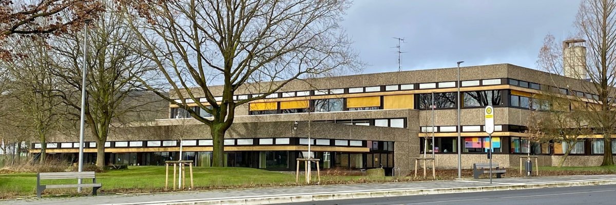 Außenansicht eines zweigeschossigen, langgestreckten Gebäudes mit dunkler Fassade und Fensterbändern. Im Vordergrund verlaufen eine Straße, ein Gehweg mit Sitzbank und eine Bushaltestelle. Mehrere kahle Bäume stehen vor dem Gebäude.