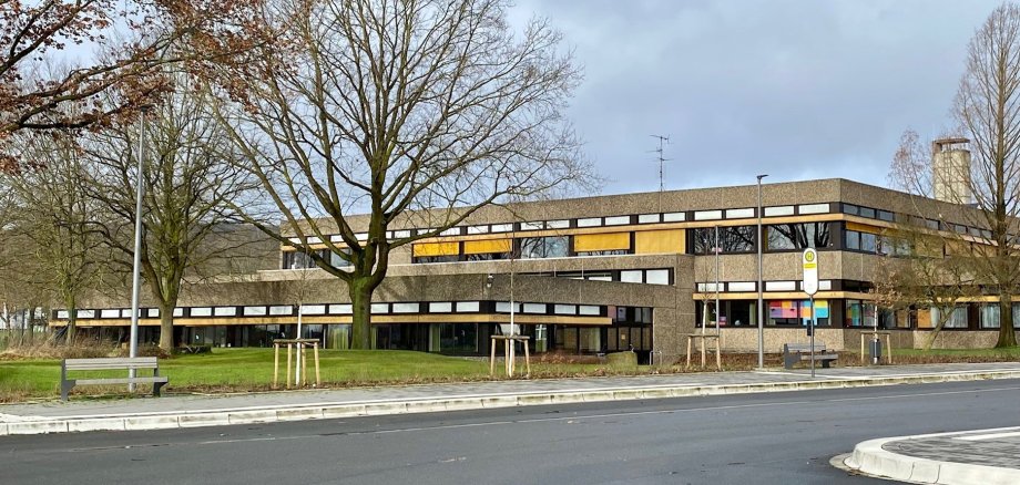 Außenansicht eines zweigeschossigen, langgestreckten Gebäudes mit dunkler Fassade und Fensterbändern. Im Vordergrund verlaufen eine Straße, ein Gehweg mit Sitzbank und eine Bushaltestelle. Mehrere kahle Bäume stehen vor dem Gebäude.
