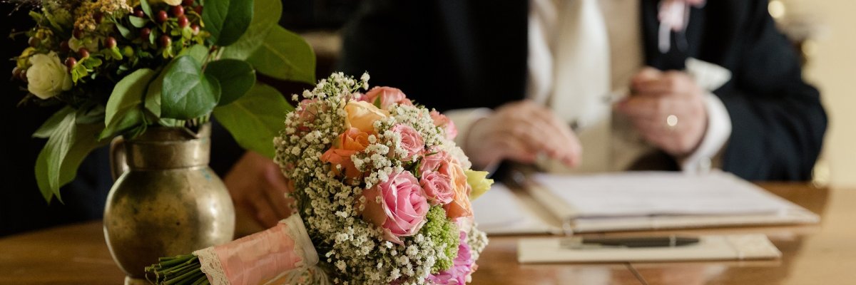 Großaufnahme eines Tisches im Standesamt, auf dem ein schöner, rosa farbender Brautstrauß liegt. Im Hintergrund unterschreibt der Bräutigam gerade die Heiratsurkunde. 