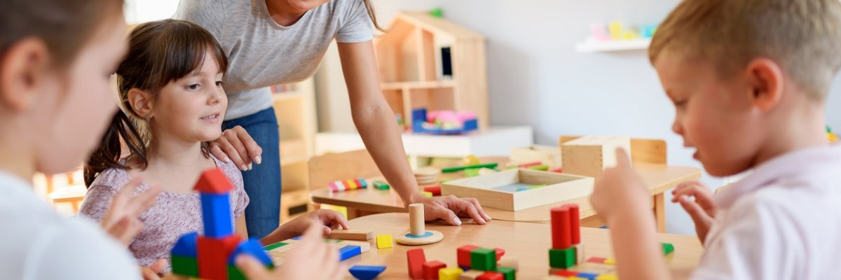 Kindertagesstätte 1 Eine Situation aus einer Kindertagesstätte. Eine Erzieherin beschäftigt sich mit mehreren Mädchen und Jungen, die an einem Tisch sitzen und mit bunten Bauklötzen spielen.