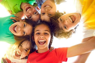 Group of happy little children playing together, having fun, huddling, looking down and smiling Sieben Kinder in bunten T-Shirts, die die Köpfe zusammenstecken und von oben in die Kamera lachen.