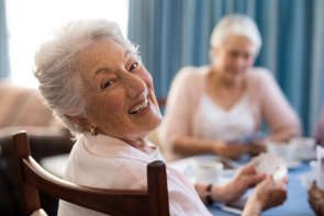 Smiling senior woman playing cards with friends Eine Rentnerin, die an einem Tisch sitzt und mit anderen Damen Karten spiel, dreht sich um, um in die Kamera zu lächeln.