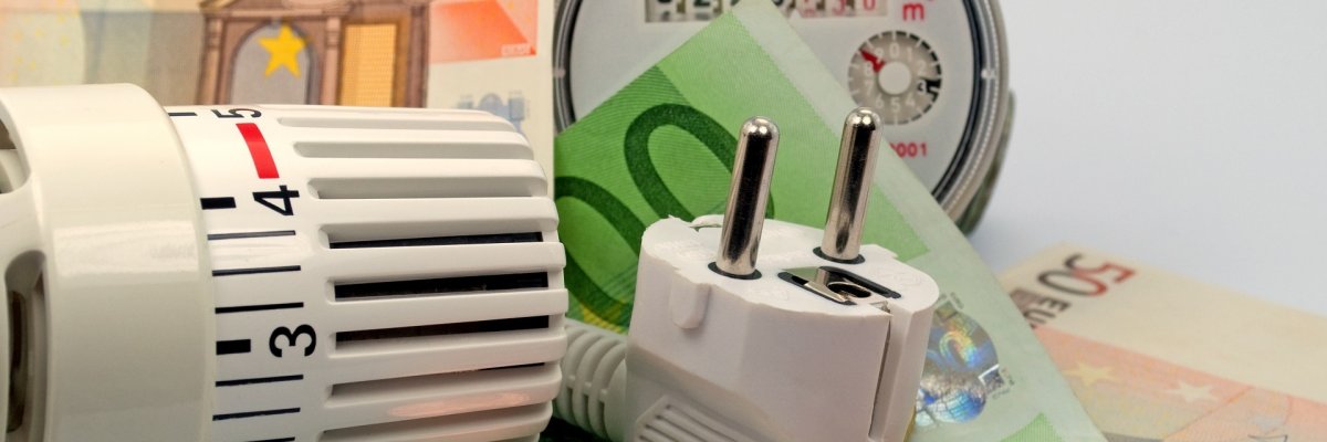 Neben einem Thermostatventil liegen Geldscheine, Geldmünzen und ein Stecker – Symbolbild für Energiekosten.