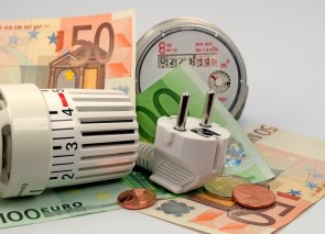 Neben einem Thermostatventil liegen Geldscheine, Geldmünzen und ein Stecker – Symbolbild für Energiekosten.