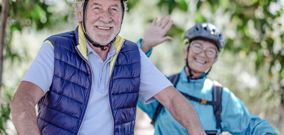 Älterer Mann mit Fahrradhelm und Fahrrad lächelt in die Kamera. Hinter ihm fährt eine weitere Person mit Fahrradhelm und Rucksack auf einem schattigen, von Bäumen gesäumten Weg.