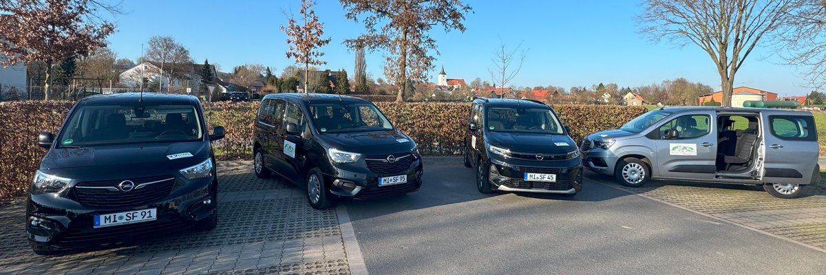 Seniorenfahrdienst Vier Fahrzeuge des Seniorenfahrdienstes stehen auf einem Parkplatz. Drei schwarze und ein graues Fahrzeug sind nebeneinander geparkt, an den Fahrzeugen sind Logos des Fahrdienstes angebracht. Beim grauen Fahrzeug ist die seitliche Schiebetür geöffnet und gibt den Blick auf den Innenraum frei. Im Hintergrund sind Bäume, Wohnhäuser und eine Kirche unter blauem Himmel zu sehen.