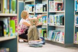 Teacher Assisting Boy In Selecting Books In Library Eine Frau hilft einem Kind, das in einem Bücherregal sucht.