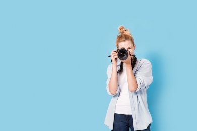 Young blonde photographer is taking a photo. Model isolated on a blue background with copy space Eine Frau die gerade ein Foto mit einer Spiegelreflexkamera macht, steht vor einer blauen Wand.