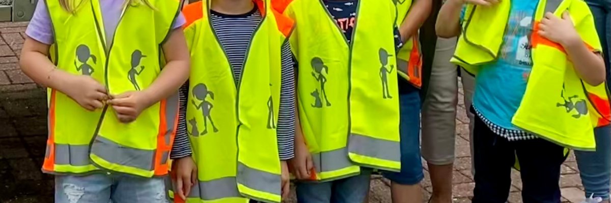 Nahaufnahme mehrerer Kinder, die im Freien nebeneinanderstehen und leuchtend gelbe Warnwesten mit orangefarbenen und reflektierenden Streifen tragen. Auf den Westen sind dunkle Figuren von Kindern abgebildet.