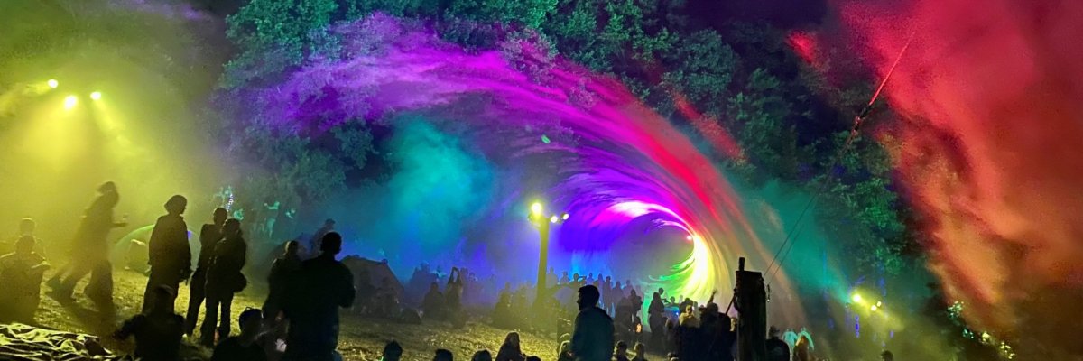 Viele Menschen sitzen und stehen nachts auf einer Wiese bei einer Veranstaltung im Freien. Im Hintergrund erzeugt eine große Lichtinstallation einen tunnelartigen Effekt aus bunten Farben wie Grün, Blau, Lila und Gelb, die sich wie eine Welle durch die Luft ziehen. Nebel oder Rauch verstärkt die leuchtenden Farben. Umgeben ist die Szene von Bäumen, die teilweise angestrahlt werden. Einige Besucher sitzen auf dem Boden oder Decken, andere stehen und beobachten die Lichtshow.