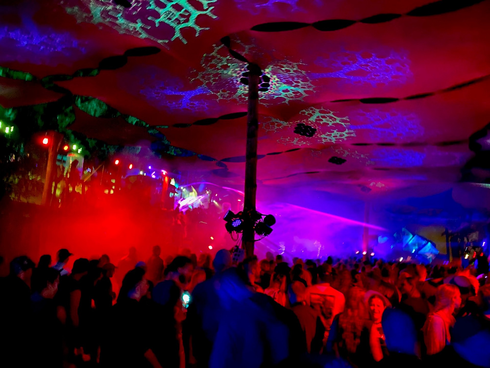 Eine große Menschenmenge steht dicht gedrängt in einer Halle oder einem Zelt und feiert bei einer Veranstaltung. Bunte Lichtstrahlen in Rot, Blau und Violett durchziehen den Raum, während Nebel oder Rauch die Atmosphäre verstärkt. In der Mitte steht eine Säule mit montierter Lichttechnik, von der aus Scheinwerfer in verschiedene Richtungen leuchten. Die Decke ist mit organisch wirkenden Mustern dekoriert. Im Hintergrund ist eine Bühne mit Lichtshow erkennbar, während im Vordergrund Menschen tanzen und miteinander sprechen.
