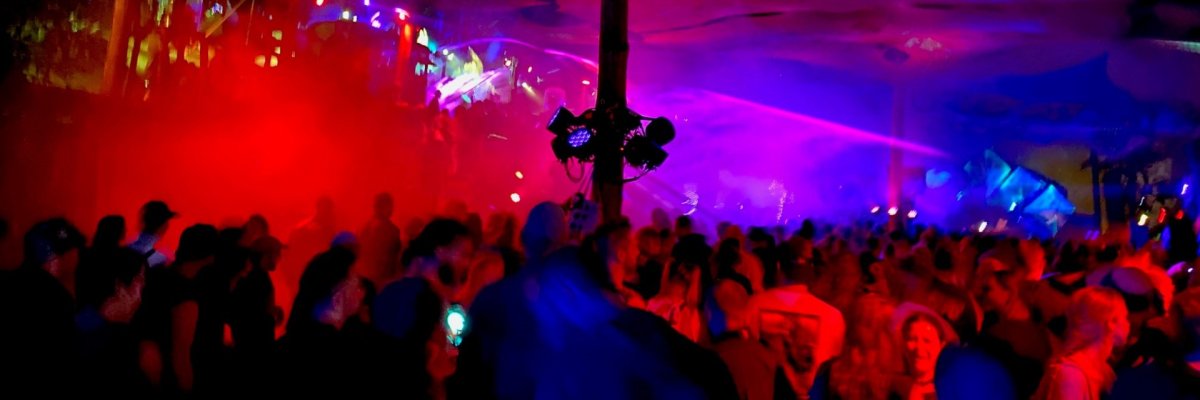 Eine große Menschenmenge steht dicht gedrängt in einer Halle oder einem Zelt und feiert bei einer Veranstaltung. Bunte Lichtstrahlen in Rot, Blau und Violett durchziehen den Raum, während Nebel oder Rauch die Atmosphäre verstärkt. In der Mitte steht eine Säule mit montierter Lichttechnik, von der aus Scheinwerfer in verschiedene Richtungen leuchten. Die Decke ist mit organisch wirkenden Mustern dekoriert. Im Hintergrund ist eine Bühne mit Lichtshow erkennbar, während im Vordergrund Menschen tanzen und miteinander sprechen.