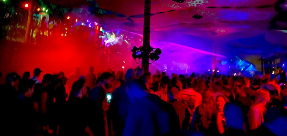 Eine große Menschenmenge steht dicht gedrängt in einer Halle oder einem Zelt und feiert bei einer Veranstaltung. Bunte Lichtstrahlen in Rot, Blau und Violett durchziehen den Raum, während Nebel oder Rauch die Atmosphäre verstärkt. In der Mitte steht eine Säule mit montierter Lichttechnik, von der aus Scheinwerfer in verschiedene Richtungen leuchten. Die Decke ist mit organisch wirkenden Mustern dekoriert. Im Hintergrund ist eine Bühne mit Lichtshow erkennbar, während im Vordergrund Menschen tanzen und miteinander sprechen.