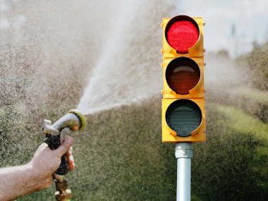 Eine rote Trinkwasserampel steht im Vordergrund. Daneben sprüht eine Person mit einem Gartenschlauch Wasser in die Luft. Das Bild symbolisiert einen hohen Wasserverbrauch und die Aufforderung, bei Wasserknappheit sparsam mit Trinkwasser umzugehen.