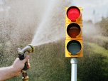 Eine rote Trinkwasserampel steht im Vordergrund. Daneben sprüht eine Person mit einem Gartenschlauch Wasser in die Luft. Das Bild symbolisiert einen hohen Wasserverbrauch und die Aufforderung, bei Wasserknappheit sparsam mit Trinkwasser umzugehen.