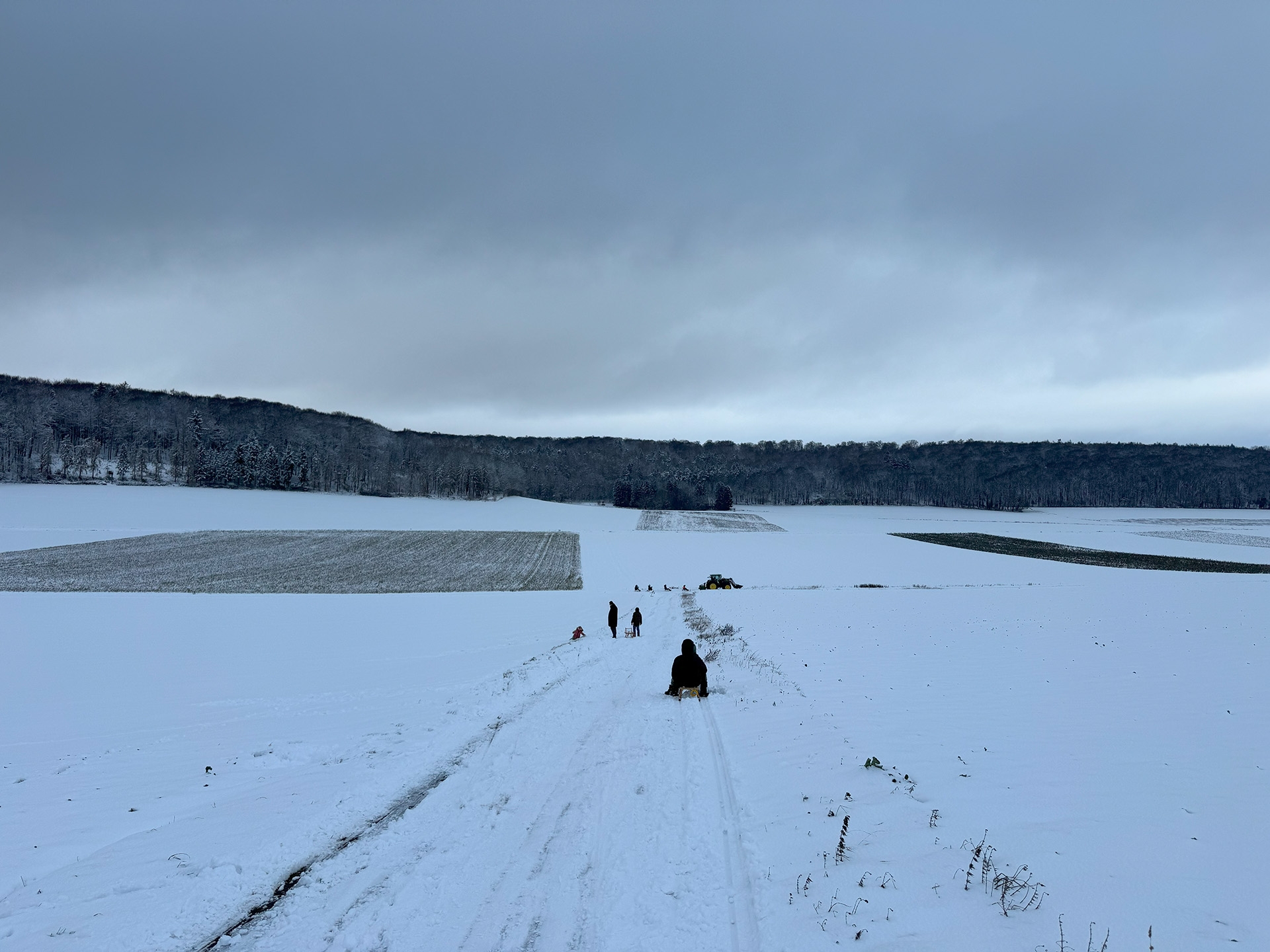 Verschneite, weite Feldlandschaft unter grauem Himmel. In der Mitte verläuft ein schmaler, festgefahrener Weg durch den Schnee. Mehrere Personen sind darauf unterwegs, einige ziehen oder fahren auf Schlitten. Im Hintergrund steht ein Traktor auf dem Feld. Dahinter erstreckt sich ein dunkler, bewaldeter Hügelrand.