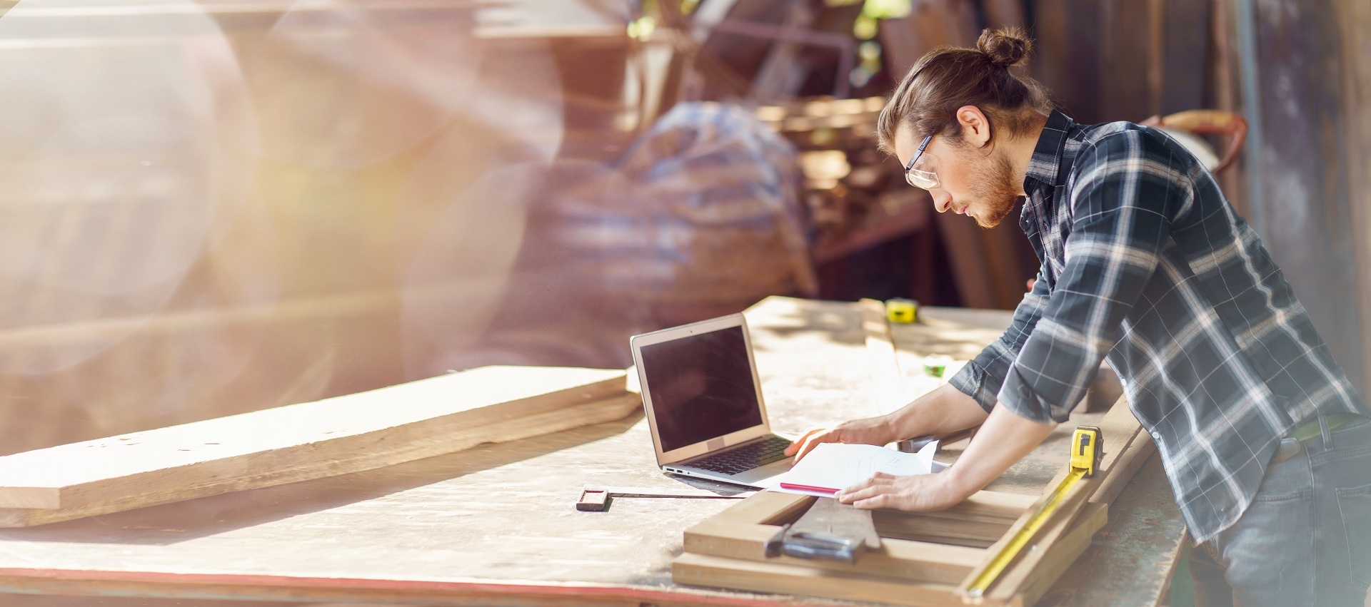 Person in einer Holzwerkstatt arbeitet an einer Werkbank mit Holzbauteilen. Neben klassischen Handwerkswerkzeugen nutzt sie einen Laptop und Unterlagen zur Planung und Vorbereitung. Die Aufnahme verbindet handwerkliche Arbeit mit digitaler Unterstützung.
