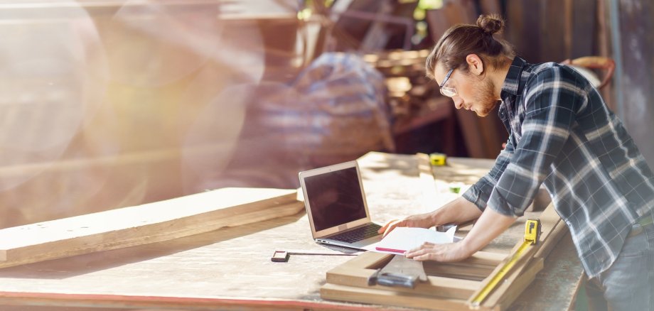 Person in einer Holzwerkstatt arbeitet an einer Werkbank mit Holzbauteilen. Neben klassischen Handwerkswerkzeugen nutzt sie einen Laptop und Unterlagen zur Planung und Vorbereitung. Die Aufnahme verbindet handwerkliche Arbeit mit digitaler Unterstützung.