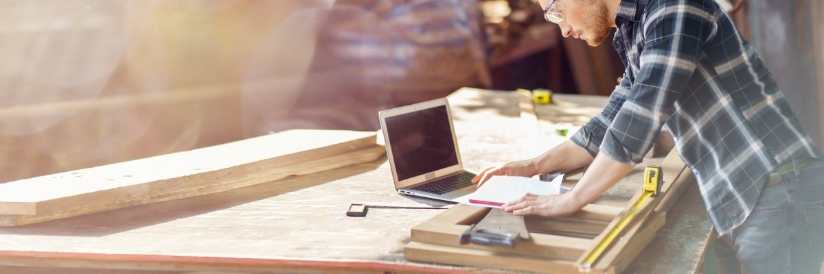 Handwerk & Digitales Person in einer Holzwerkstatt arbeitet an einer Werkbank mit Holzbauteilen. Neben klassischen Handwerkswerkzeugen nutzt sie einen Laptop und Unterlagen zur Planung und Vorbereitung. Die Aufnahme verbindet handwerkliche Arbeit mit digitaler Unterstützung.