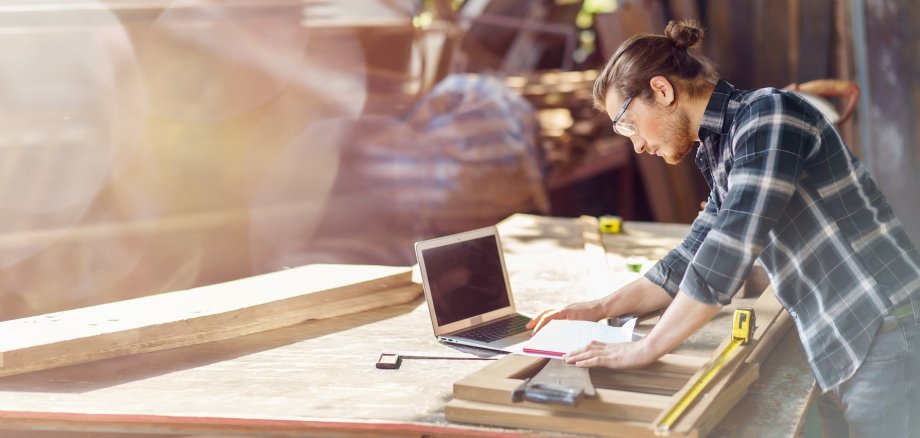 Handwerk & Digitales Person in einer Holzwerkstatt arbeitet an einer Werkbank mit Holzbauteilen. Neben klassischen Handwerkswerkzeugen nutzt sie einen Laptop und Unterlagen zur Planung und Vorbereitung. Die Aufnahme verbindet handwerkliche Arbeit mit digitaler Unterstützung.