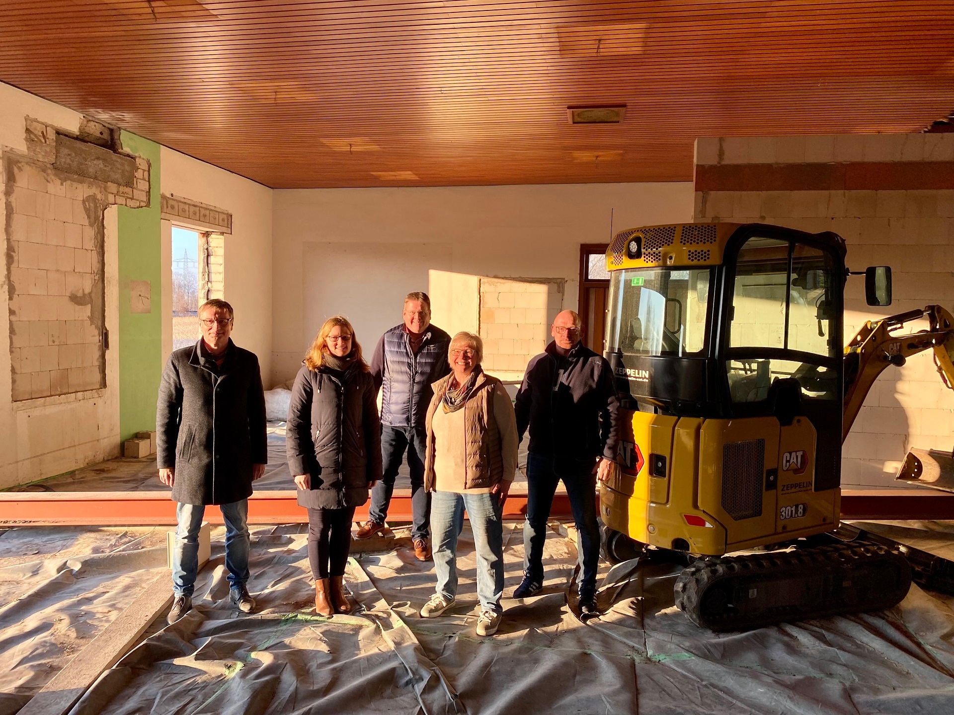 Mehrere Personen stehen neben einem gelben Bagger auf einer Baustelle - einem Saal, der gerade umgebaut wird. 